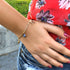A close-up of a person in a floral red top and denim shorts, with their hand on their hip, showcases them wearing the Women Mega Lucky Charms Bracelet. This 14k gold-filled piece is adorned with five meaningful amulets, including a small blue evil eye charm, designed to bring positive vibes. The lush greenery in the background enhances the serene aura of the scene.